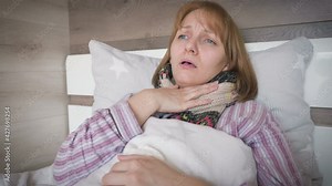 Beautiful sick woman lying on bed, suffering from flu or coronavirus symptoms. Adult ill female coughing and taking a pill with water as having a temperature.