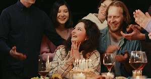 Woman blowing out candles on birthday cake surrounded by clapping friends