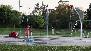 Child playing with fountains in local public park. Splash pad and swings at playground on summer day.