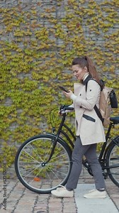 Full length portrait of elegant woman in beige trench coat with bicycle and backpack sending text message using smartphone against the green wall outside in the city.