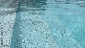 Close-up summer rain falls in large drops into a blue clear swimming pool in hotel and leaves round reflections on the water