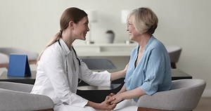 Happy positive doctor woman giving support to senior patient, telling optimistic, positive prognosis, holding hands, touching shoulder, sitting in armchair, speaking, smiling