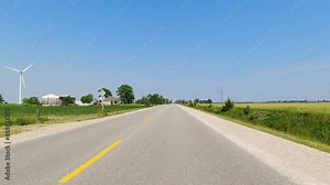 Front view Driving Plate looking forward. POV driving car view. Rural straight road with turns right side road. Summer daylight farm fields building and windmill. Road ahead no cars.