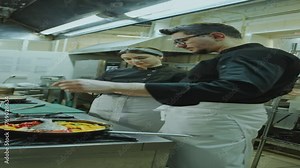 Restaurant chef explaining how to cook fish with vegetables to young female apprentice taking notes in copybook during internship in the kitchen