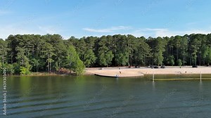 sideways aerial footage of a sandy beach along the banks of Lake Acworth with rippling green waters and people in the water and relaxing on the beach with umbrellas in Acworth Georgia USA