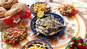 Traditional Moroccan summer food: grilled sardines, fresh vegetable salad, Moroccan bread and fruits. a man's hand puts a lemon next to sardines.
