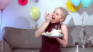 Woman in festive dress and party cap eating birthday cake with her hands, her face stained cream. A woman stuffing her face with cake. Celebrating birthday party at decorated home. Selective focus.