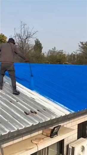 spraying bright blue paint onto the grey corrugated metal roof using a long handled power sprayer