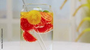 Fresh red and yellow cherry tomatoes and basil in sparkling water in glass jar with straw rotating on white table on light background close up. Detox drink, tomato juice, vegetable healthy drink bever