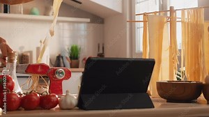 A woman preparing pasta using digital tablet, checking online recipe in tablet using mobile cookbook menu apps in sunny kitchen. A woman making fresh fettuccine using pasta machine