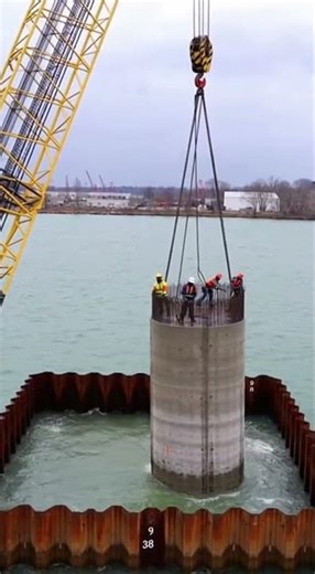 A barge crane lowers a heavy circular pillar into a rushing river while workers balance on top Wate