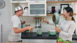 Asian Young couple using kitchenware utensils as helmets and swords playing fight battle in kitchen activity at home. They laugh with happiness. Family relationship concept