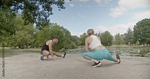 A fit sports couple stretches and warms up outdoors in a beautiful park by the lake on a bright, sunny day, preparing for their workout.