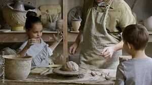 PAN of male potter wearing apron putting piece of clay on pottery wheel and wetting it with sponge while teaching curious children in his workshop