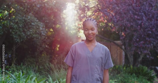 Woman wearing grey scrubs responding to sun shaft, tilting head and crossing arms in garden