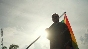 LGBTQ gay Man standing proudly, holding and raising the rainbow LGBTQ+ flag against a clear blue sky with sunlight. LGBTQ proud celebrating Pride Month and the ongoing fight for equality and love.