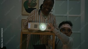 Low key shot of cheerful African American mother and her little son setting projector to watch movies at home