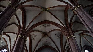Beautiful stone arches on the ceiling of a gothic cathedral with few people sitting and praying on the hard wood benches underneath. Smooth downwards tilt