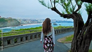 Young woman walking along the seashore in Biarritz, France. Beautiful girl in a dress looking at the Biaritts city by the cliffs.