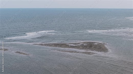 Drone footage captures a wide sandbar exposed during low tide, with gentle waves lapping around the formation and open ocean in the background.