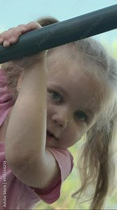 VERTICAL VIDEO: Little girl climbs gymnastic ladder on open sports ground on outside. Close up portrait of cute little girl crawls on vertical sports ladder in city park on sun day. slow motion