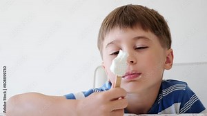 Handsome boy eating popsicle on a stick. He bites off the ice cream with pleasure. Close-up. Child eat in the kitchen or at the dining room table. Tasty dessert. Slow motion. High quality 4k footage