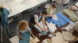 Team of professional dressmakers chalking sewing patterns on fabric and having discussion, working together on clothing at table in sewing workshop. High angle view