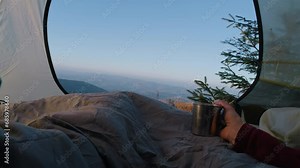 Romantic hiker couple lie in tent on mountain hill and drink hot tea. Two tourists rest under blanket during adventure vacation. Backpacker family warm up at cold windy weather and admire the scenery.