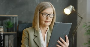 Caucasian businesswoman holding and using tablet device in hands. Beautiful senior woman in glasses tapping and scrolling on computer in office. Female texting and typing and thinking in cabinet.