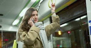 Young pretty happy woman riding bus talking on phone smiling. Attractive adult stylish caucasian female standing in empty public transport holding handrail enjoying ride wearing coat and scarf.