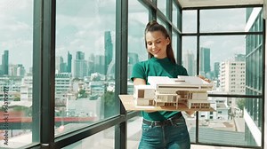 Young happy beautiful engineer check house model by turning left side and right side. Professional manager inspect, lifting, looking at architectural model while standing near window. Tracery
