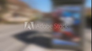 A bustling urban intersection captured in a single shot, featuring a bus stop located alongside a parked car on a busy street. The defocused view adds depth to the dynamic city scene
