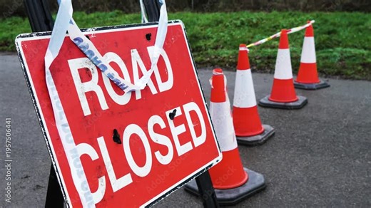 Road Closed sign standing near restricted roadway. Traffic cones blocking access while warning tape stretching across entrance. Temporary prohibition preventing vehicle passage and restricting traffic