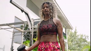Fit biethnic woman trains with dumbbells in posh tropical gym. Female athlete builds biceps, embodies strength, vitality outdoors. Health-focused active lifestyle routine in an exotic, lush setting.