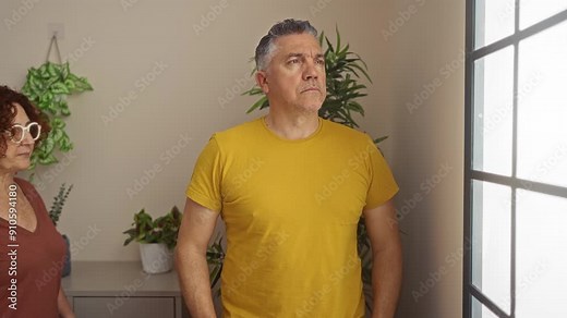 Middle-aged man and woman standing together in a living room indoors, sharing an intimate and caring moment while looking out a window