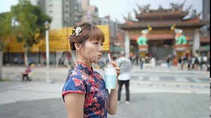 A young Taiwanese woman in a blue traditional cheongsam dress sips bubble tea while walking past historic buildings in Wanhua District, Taipei City, Taiwan. slowmotion