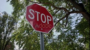 Handheld slow motion shot rotating around red stop sign by street sidewalk near public park with green tree branches and nature in the background