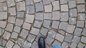 Women's feet walk along a cobblestone path, first-person view. Close-up of human feet walking down the street. A walk on a sunny day in spring or autumn. Top view of historic stone cobblestone street