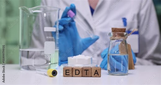 Wooden blocks spell abbreviation EDTA near lab glassware and corked vial. Gloved researcher dispenses solution with pipette measuring sample in beaker