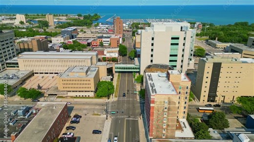 An aerial perspective showcases the vibrant downtown area of Waukegan, Illinois, featuring various buildings, roads, and a pedestrian bridge, all leading towards the picturesque blue waters of Lake