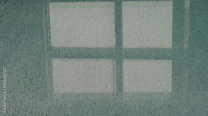 Raindrops on a window pane create a soothing pattern, with the shadow of a grid visible in the background. The scene evokes a calm and rainy atmosphere.
