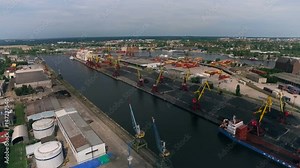 Sea Port. Cargo Ship in Dock. Cranes Loading Coal. Storage Houses on Shore. Containers on Ground. City Buildings on Background. Green Trees. Summer