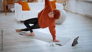 Fit slim african american young girl in sportswear doing side plank exercise at home on mat. Curly redhead woman training online using tablet. Workout yoga practice fitness online exercising concept.