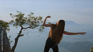 Back view Slender young woman standing on mountain among rocks overlooking ocean and islands, arms outstretched in different directions. Back view woman doing fitness outdoors on a sunny day