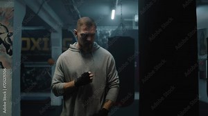 Athletic boxer stands in dark boxing gym and looks at camera. Male fighter in boxing bandages stretches, hits punching bag and prepares for training. Physical activity and intensive workout.