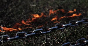 Galvanized rope battle chains for sport exercises lie on burning black grass. Durable metal chains lie in fire on lawn slow motion