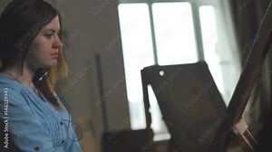 Drawing to music. A female artist paints a painting on canvas, listening to music on headphones. Classical music and fine arts. A young woman is drawing behind an easel. Stock Video