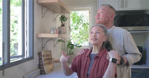 Senior Asian couple by large kitchen window, noticing movement outside, pointing and smiling