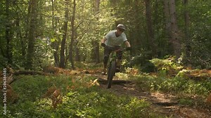 SLOW MOTION: Extreme downhill bike racer rides over a log on the dirt road leading through the sunlit forest. Young man having fun speeding through the forest on his bright green electric bicycle.