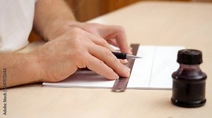 Designers hands drawing lines on white blank with iron ruler and brush pen. Close shot. Side view. Preparing blank for calligraphic lettering. Inkpot with black paint near hands. Blurred background